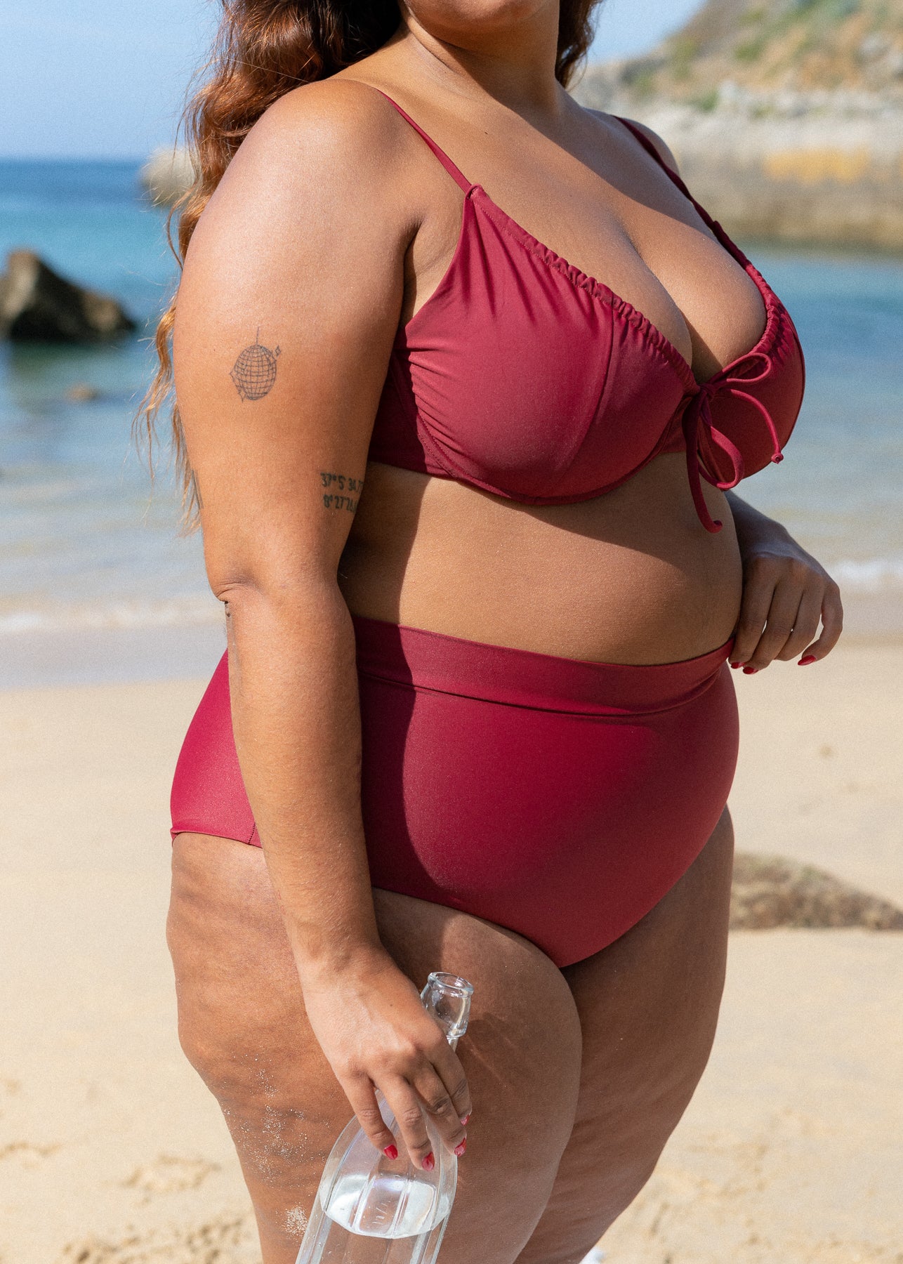 Red drawstring bikini top by Lilja the Label with tie detail, worn with high-waisted bottoms on the beach.