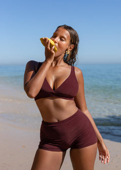 Burgundy mini shorts by Lilja the Label, paired with a matching bikini top, on a model eating fruit at the beach.