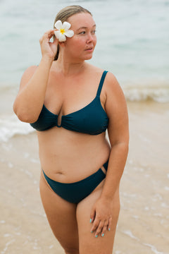 Teal high-waisted swim bottoms with side cutouts, worn by model on beach with white flower in hair, side view.